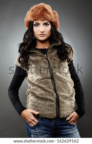 Portrait of an attractive young woman wearing fur vest and cap with hands in pockets over gray background