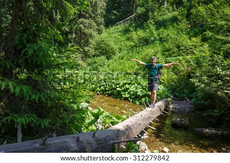 Happy male hiker crossing a mountain river on a huge tree trunk