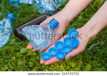 Hand of woman with plastic bottle and bottle caps, rubbish on grass in background, concept of environmental protection, littering of environment