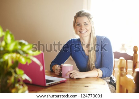 Young woman sitting at table with laptop and hot drink smiling