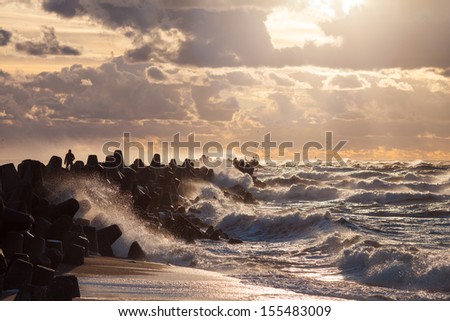 Silhouette of a man is facing storm standing on water breaker with waves and water splashes on Baltic sea while sunset