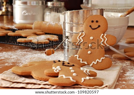 Smiling gingerbread man standing next to flour sifter with baking ingredients and additional gingerbread cookies in background.  Partially decorated cookies in foreground.  Closeup with shallow dof.