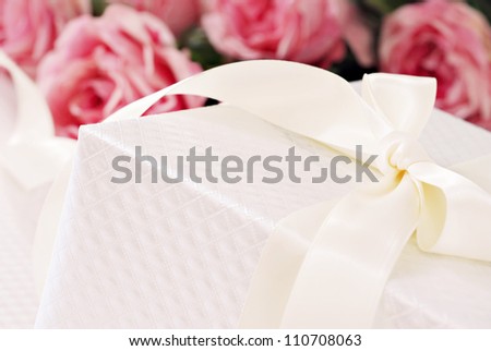 Elegant cream colored gift box with satin ribbon and bouquet of roses in background.  Macro with shallow dof and copy space.  Selective focus on corner of box and bow.