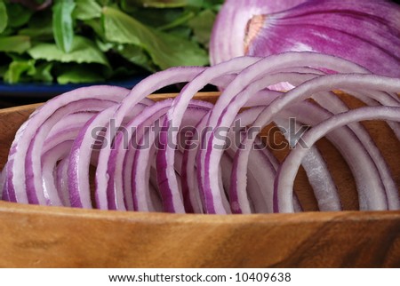Red onion rings in a wooden bowl with whole onions and salad greens in the background.  Close-up with focus on the rings and shallow dof.