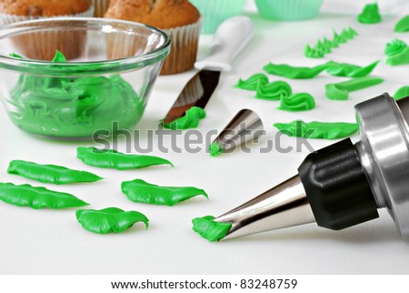 Green cake frosting being dispensed from decorator tip to form leaves.  Bowl of frosting and cupcakes in background along with practice elements of cake decorating.  Macro with shallow dof.