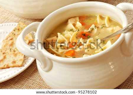 Chicken noodle soup in cream colored ceramic bowl with handles.  Plate of crackers and soup tureen in background.  Closeup with shallow dof.