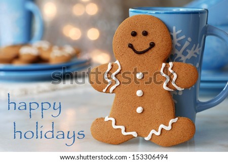 Holiday greeting card with smiling gingerbread man standing next to snowflake mug.  Plate of additional cookies and defocused holiday lights in background.