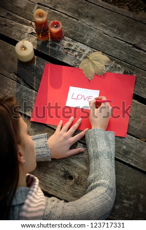 top view over the shoulder of a woman writing word LOVE on white and red paper on wooden table with candles and leaf