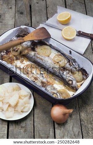 Baked mackerel fish in casserole with wooden scoop on old wooden table with whole onion, cut lemon and potato salad on gray wooden background.