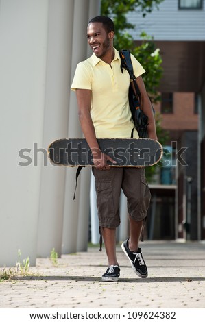 Happy young African American male college student holding his skateboard and walking on campus