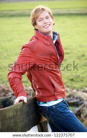 Casual young blonde guy smiling and looking over his shoulder. He is in a relaxed pose and leaning against a wooden fence outdoors.