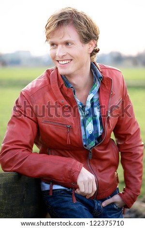 Handsome young blonde male relaxing outdoors. Casual guy wearing a red leather jacket and blue jeans and shirt. He is leaning against a fence outdoors and smiling while looking away.