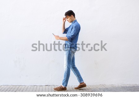 Full body portrait of a young man walking and reading text message on cell phone