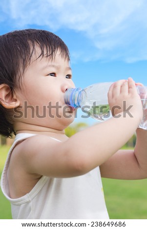Little asian girl  drinking water from plastic bottle, after tired from a romp in the park, under bright sunlight and blue sky background, outdoor shot