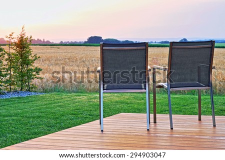 Two outdoor chairs on a wooden veranda, in front of a field and the sunset.