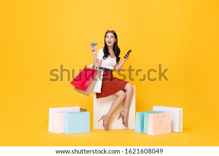 Beautiful Asian woman sitting and carrying shopping bags with credit card and mobile phone in hands on yellow background
