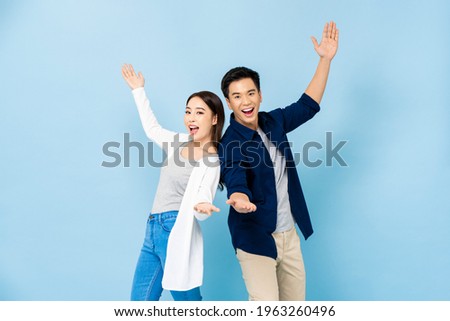 Surprised Asian couple standing back to back smiling and opening hands in light blue isolated studio background