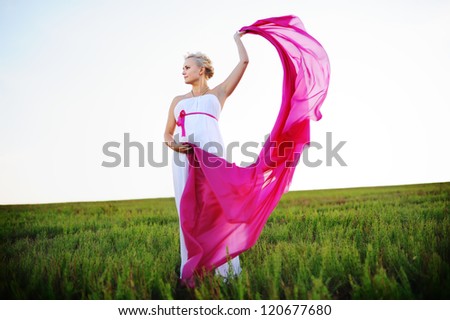 young woman in a white greek dress stands in field holds pink cloth