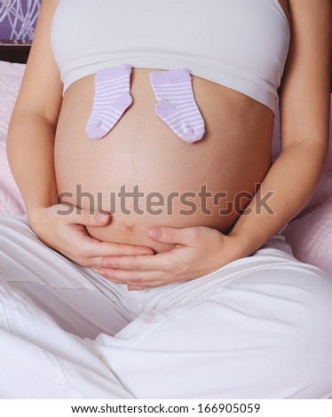 young pregnant woman sitting on bed at home, holding tiny socks for a baby