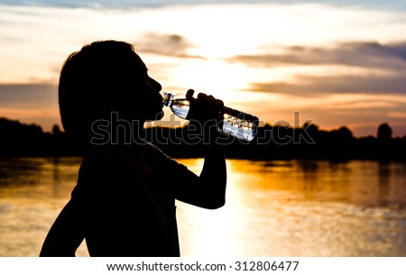 Silhouette of woman is drinking water during exercise over beautiful sunset background.