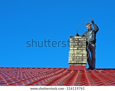 NICA, LATVIA - SEPTEMBER 25, 2013: Young man in black dresses is standing on red roof and cleaning chimney with metal brush on long cable.