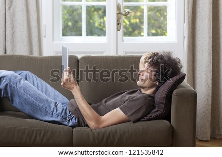 A man reclines on a sofa reading from a tablet computer.