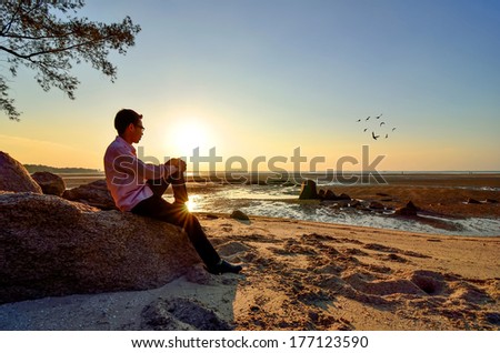 A lonely man sitting on the rock near the beach when the sun goes down.