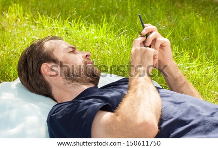 Forty years old caucasian man looking at mobile phone while laying on grass in park during a sunny summer day