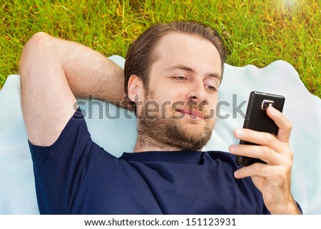 Happy smiling forty years old caucasian man looking at mobile phone while laying on grass in park during a sunny summer day