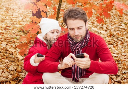 Happy smiling father and daughter looking at the mobile phone while sitting outdoor in an autumn park - enjoying modern technology