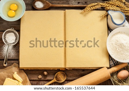 Rural vintage wood kitchen table with blank cook book, baking cake ingredients (eggs, flour, milk, butter, sugar) and cooking utensils around. Country background with free recipe text space.