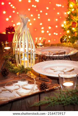 Christmas table setting. Elegant white plates with cookies, natural pine tree branch, pinecones, candles, lights and gifts on vintage planked wood. Rustic style decorations on red bokeh background.