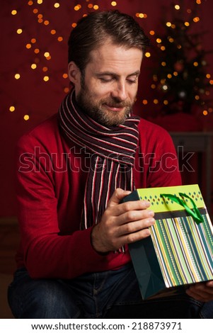Christmas - happy smiling forty years old caucasian man looking inside gift bag on dark red background with lights.