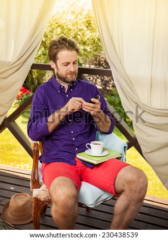 Happy forty years old caucasian man looking at mobile phone outdoor on garden terrace during sunny summer day. Countryside weekend or holiday concept.