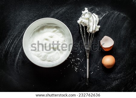 Whipping an egg whites into a foam recipe - composition on black chalkboard from above. Simple modern kitchen poster design with white bowl, eggbeater and eggshell.