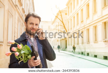 Happy smiling forty years old caucasian man with wine and flower bouquet talking on a mobile phone. Street and city buildings as background.