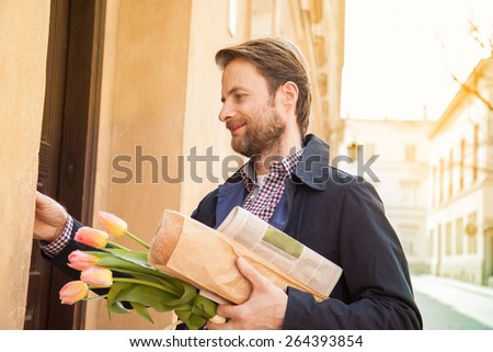 Happy smiling forty years old caucasian man with baguette, newspaper and flower bouquet ringing doorbell. Street and city buildings as background.