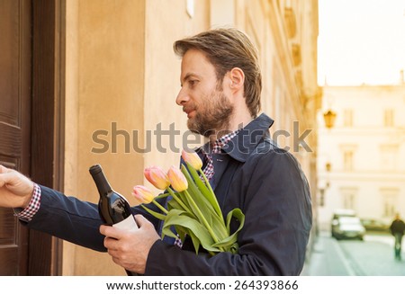 Forty years old caucasian man with wine and flower bouquet ringing doorbell. Date or celebration visit concept. Street and city buildings as background.