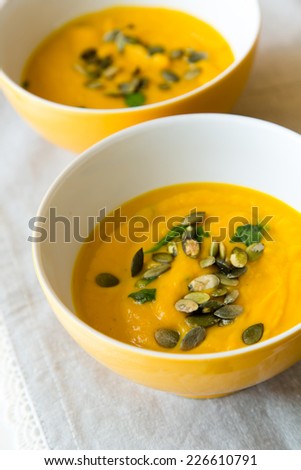 Two china bowls of homemade pumpkin soup with pumpkin seeds and herbs on laced table mat