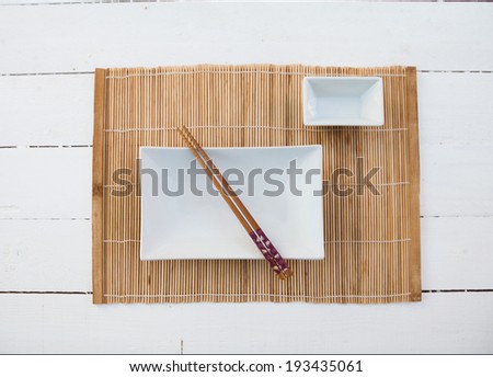 Two white squared plates with chopsticks and a bamboo table cover on a white wooden table