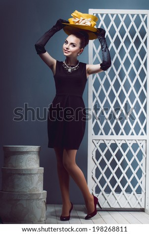 Young girl  make up in a little short black dress tries to head the yellow hat, black long gloves, hat boxes in studio.