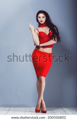 sexy brunette girl in a red skirt and red corset and red shoes with red garter stands near gray wall and wooden floor in a studio  pin up