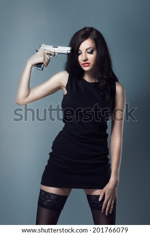 sexy brunette girl with makeup and red lips in black dress and stockings boots holding a gun to his temple