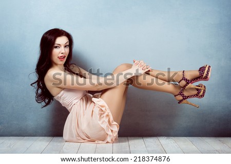 Sexy young beautiful brunette with long hair, with a slender figure sitting on lifting legs up showing tongue, licking her lips in a pink dress and make-up  retro woman athletic and acrobatic pin up