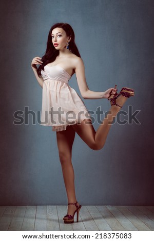 Sexy young beautiful brunette with long hair, with a slender figure standing on a lifting leg up holding his heel in a pink dress andretro woman athletic and acrobatic pin up  make-up