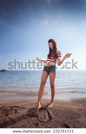 pin up young beautiful girl on the sea in short jeans and striped T-shirt with long hair rocks glasses gulls water clouds, day, wave, dream romance draws a finger in the sand heart
