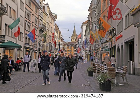 ZURICH, SWITZERLAND - MAY 04, 2013: People on cobbled pedestrian street Rennweg with boutique shops, flags on old buildings and St. Peter church clock tower in Zurich, Switzerland.