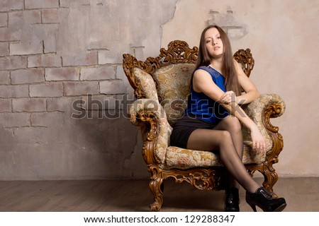 Attractive woman in a short blue miniskirt posing in an antique armchair against a grunge neutral stone wall
