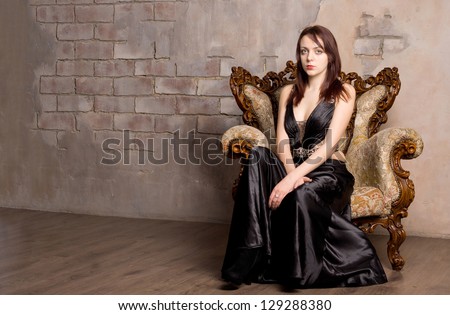 Glamorous woman in an evening gown posing for her poirtrait seated on a vintage wooden armchair against an old stone interior wall