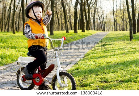Proud little boy boy with his bike standing on a paved path through a wooded park pointing his finger into the distance